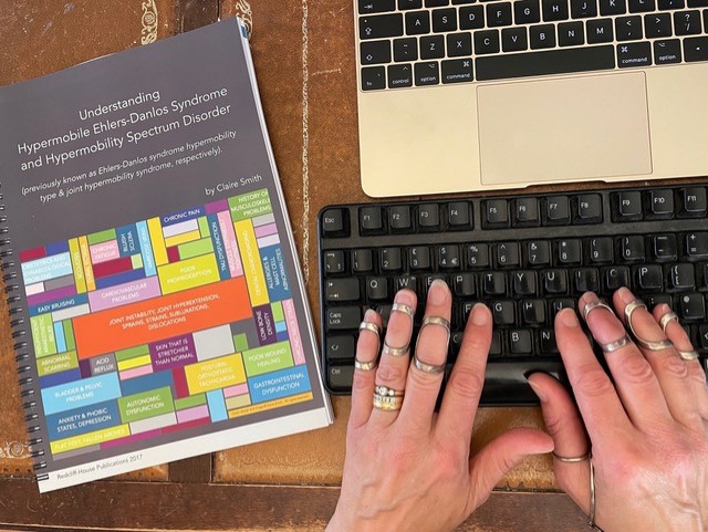 Hands typing at a computer keyboard with author tools: silver ring splints to prevent hypermobile finger joints from dislocating, second keyboard for eye-height monitor (to keep neck joints aligned), and a copy of Understanding Hypermobile Ehlers-Danlos Syndrome and Hypermobility Spectrum Disorder, by Claire Smith.