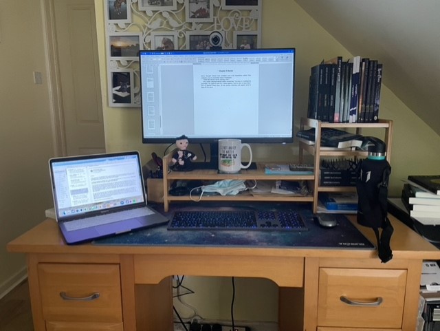 Buchanan's slightly cluttered desk, tucked into the space under the stairs. There is a flat screen monitor docked with a laptop, and a crocheted Crazie Coppa policeman propping up the monitor. A mug with the legend, "Do not annoy the writer — she might put you in a book and kill you" sits on the raised stand for the monitor and there are books on the shelf to the right of the picture, a selection of books that Kerry has written, edited, or contributed to. Behind the monitor is a framed photo montage showing family and pets and there is a bottle of water close to hand as writing can be thirsty work! A couple of masks and a packet of lateral flow tests complete the picture.