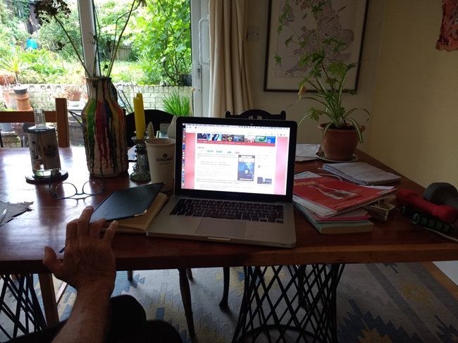 Photograph of David Thorpe's worktable in front of the French window leading onto the garden. My open laptop is displaying a page from my website about my YA science fiction novel Hybrids. The latest copy of New Scientist magazine is on the table with my paper diary and a cup of water, a vase of flowers, houseplants. notebook and exercise aids. On the wall is a map of the British Isles displaying where famous writers have lived.