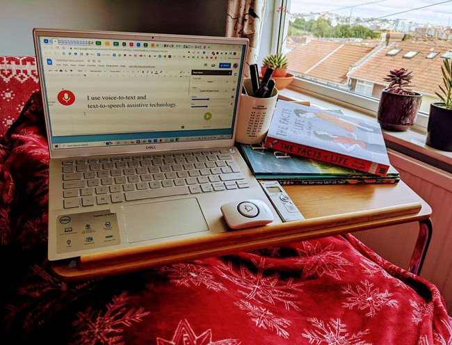 A wooden over-bed trolley holding: Books, a Scrabble mug with ink pens; an open silver laptop, dictaphone; small alarm bell unit. The document open on the laptop shows the text 'I use voice to text and text to speech assistive technology', and it shows the assistive technology icons. The bed is covered in a red blanket with white snowflake pattern. There are succulent plants on the windowsill with a view of Bristol in the background.
