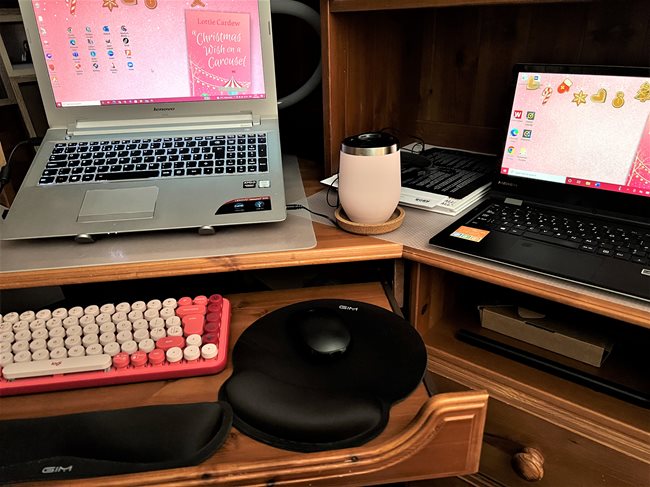 Dark wood corner desk with two open laptops. A pink typewriter-style keyboard and a black mouse, with wrist rests for both, are on a slide-out section of the desk. There is a pink thermal mug and some paperwork between the laptops, one of which is raised at an angle so the screen is at eye-level. The screensaver features an image of A Christmas Wish on a Carousel by Lottie Cardew.