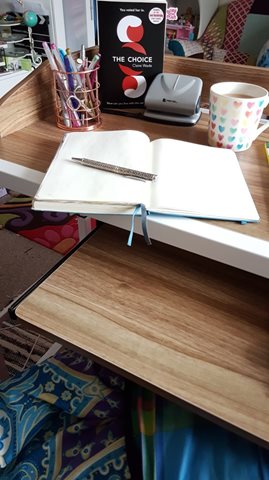An over-bed desk with author tools: an open notebook, pen, pen pot, hole punch, cup of tea and a copy of The Choice by Claire Wade.