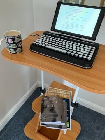 A small, height adjustable desk with bluetooth keyboard and tablet, cup of coffee and copies of ‘Where Does it Hurt?’ by Tim Atkinson plus the magazine “Pain Matters”.