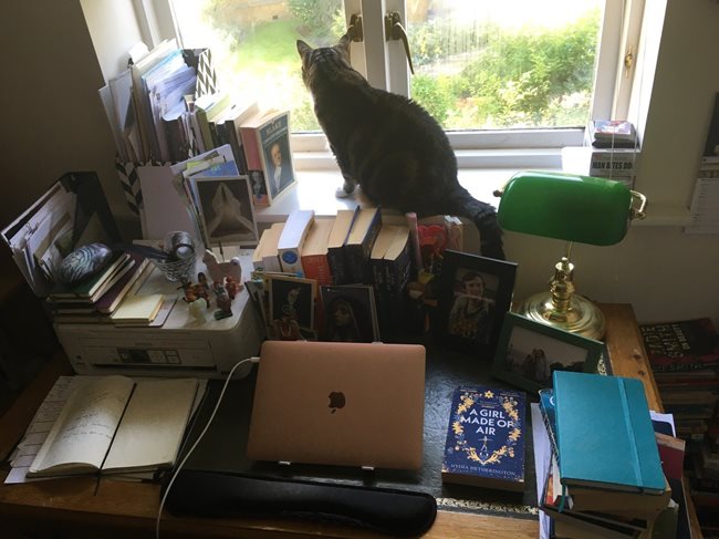 A classic pedestal desk with laptop, notebooks, small lego figures of: David Bowie in his many characters & William Blake standing atop a printer, along with other notebooks, postcards (mainly of angels) and a feather in a junk pot. There are two framed photos, a classic desk lamp, a few books stacked and piled, including A Girl Made of Air by Nydia Hetherington. Behind the desk is a window with a few more books sitting on the window sill, and a cat.