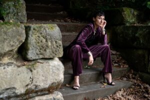 Brandy Schillace, a white woman with dark hair in a purple suit, is sitting on some stone steps.