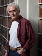 Man with grey hair wearing white t-shirt and red shirt, staning against bookcases looking into camera