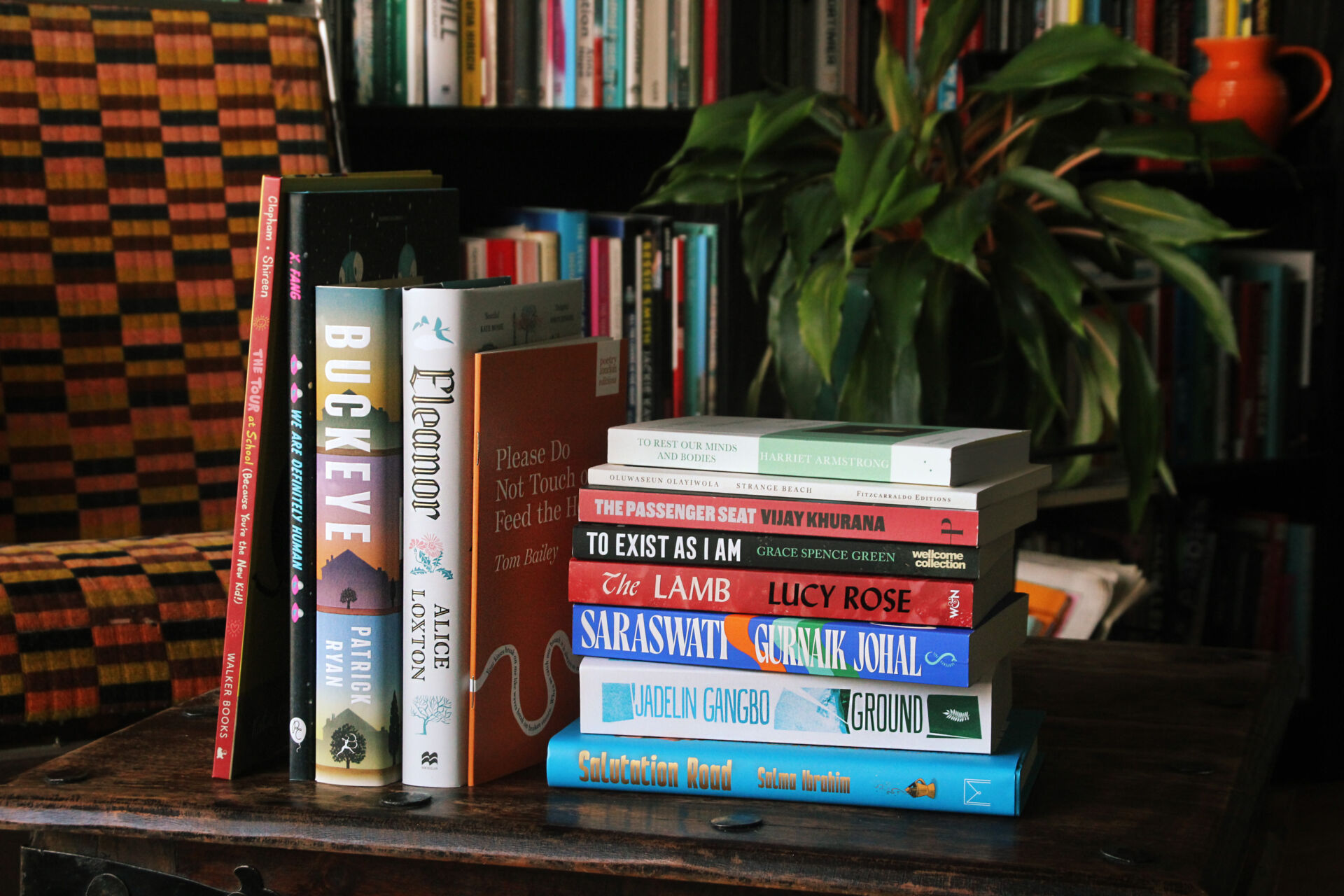 A stack of books set against a plant and a bookshelf