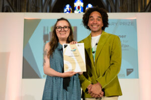 A smiling man and woman together on a stage. The woman is holding up a certificate.