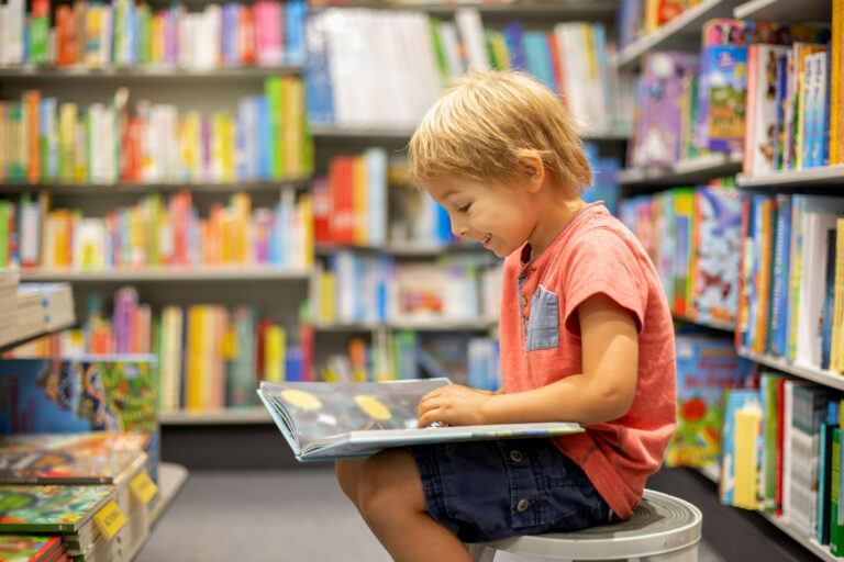 young boy happily reading, surrounded by books in a library or book shop.