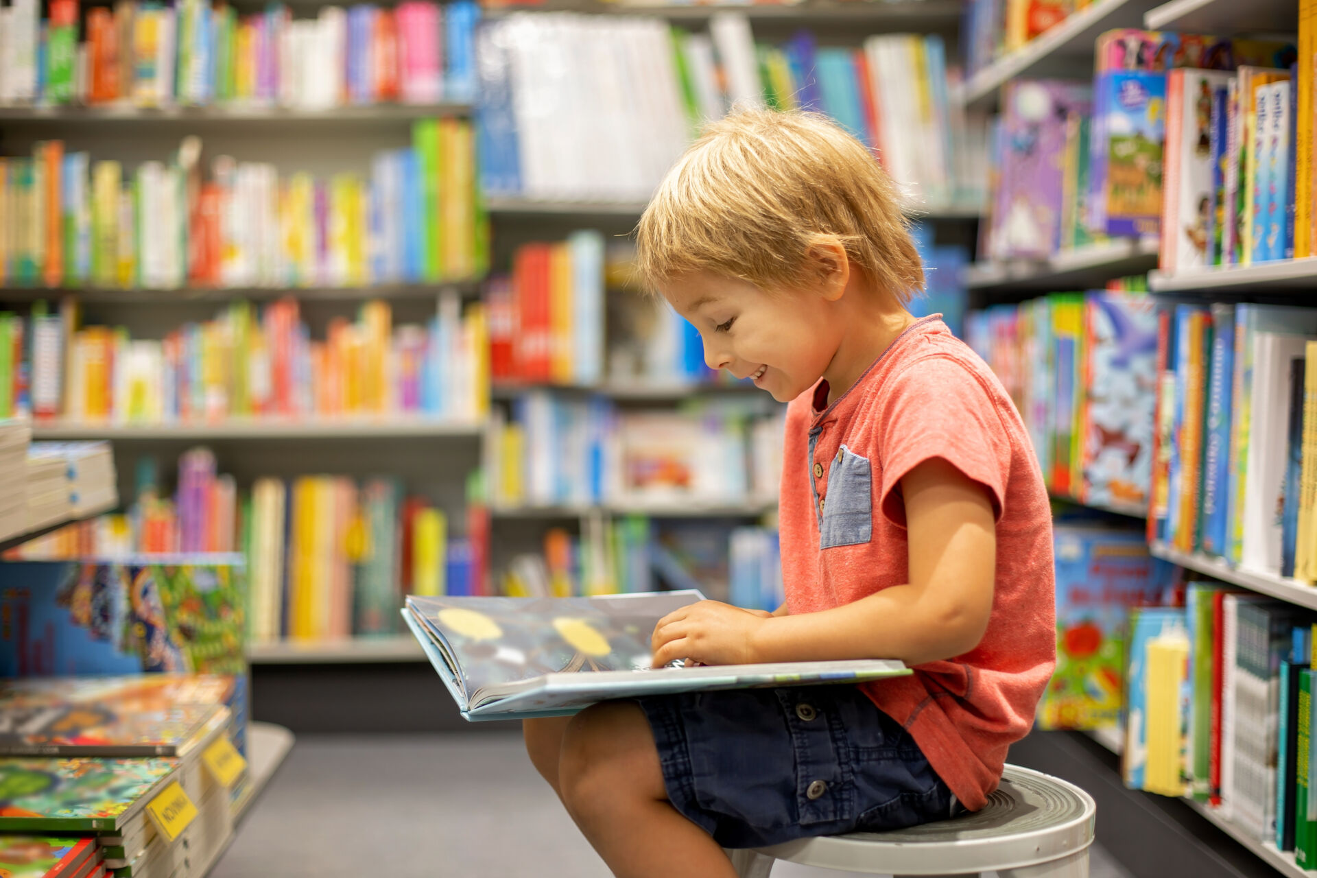 young boy happily reading, surrounded by books in a library or book shop.