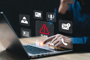 A person on a laptop with a number of icons floating in the air and red warning triangle with the word Scam in the middle