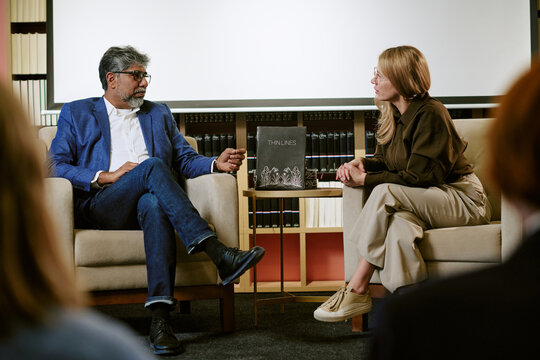 Middle aged Caucasian woman and middle aged South Asian man sitting in chairs discussing book during presentation in front of bookshelf and projection screen