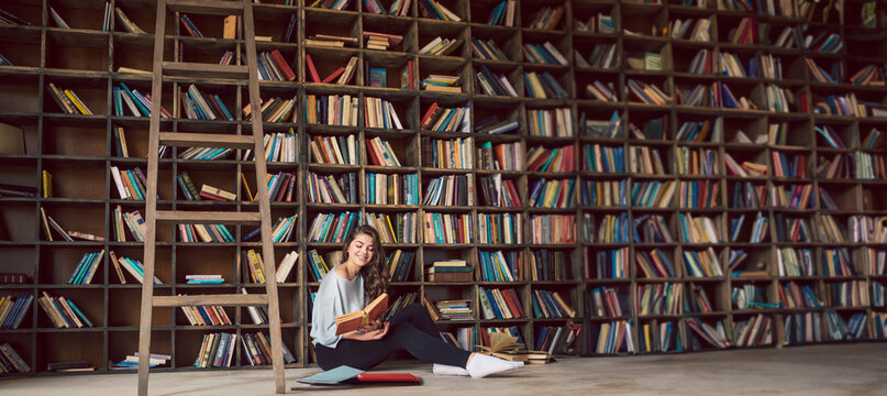 girl sitting on the floor in big library with many bookshelves at the background.