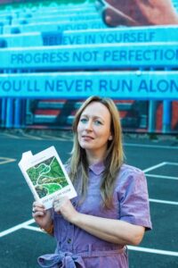 Poet Michelle Heighway, a white woman with blonde hair and blue eyeliner in a purple dress, is holding a book whilst standing in front of inspirational slogans.