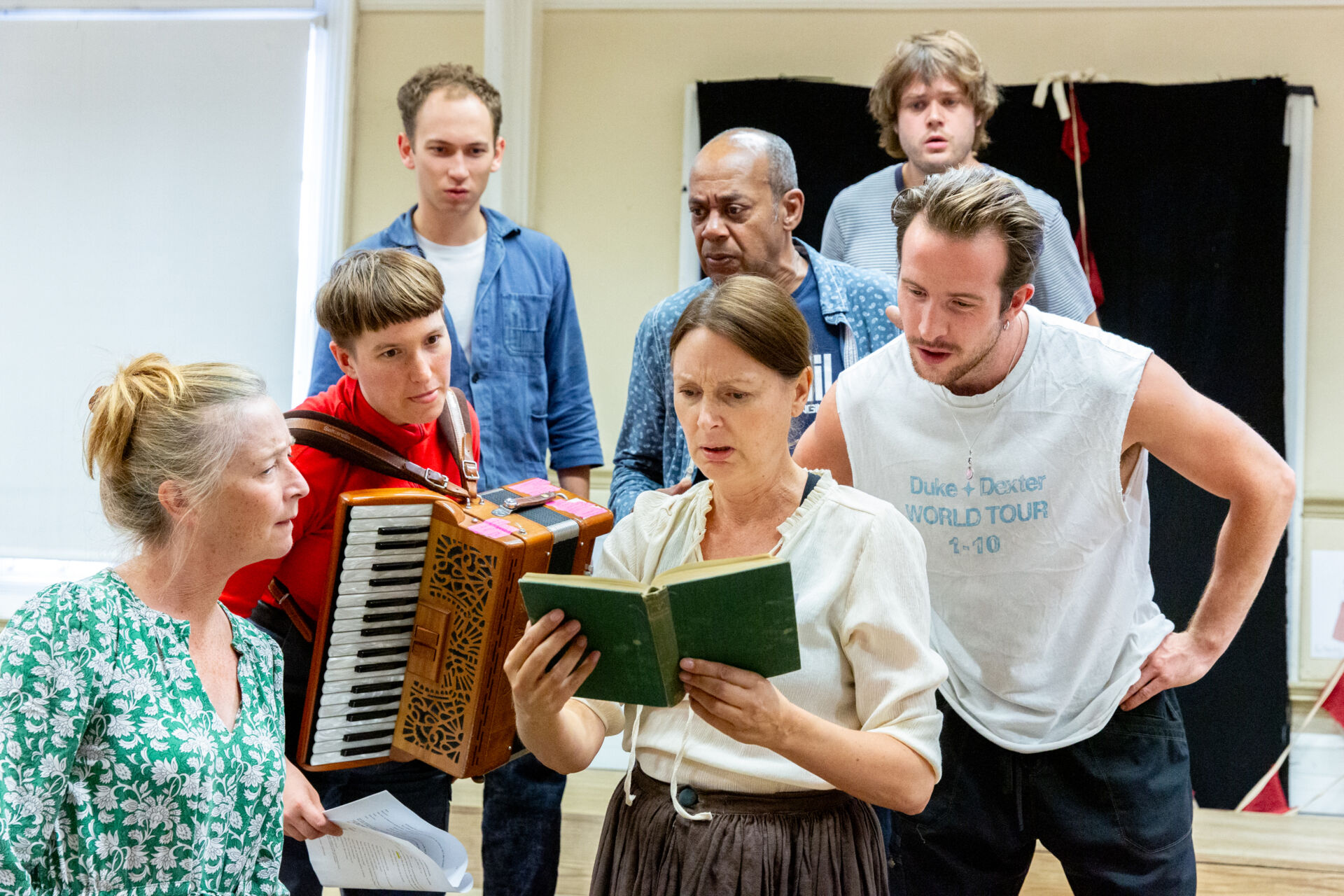 A group of actors rehearsing a play, one has an accordian, another is holding a book and all the others are looking over her shoulder. They look confused.