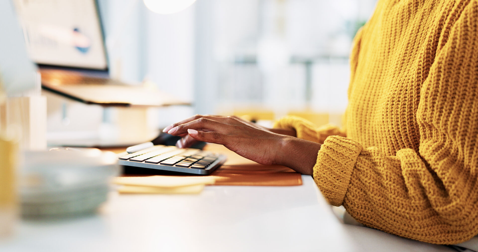 A person in a yellow jumper sitting at a laptop, you can just see their arms and hands.