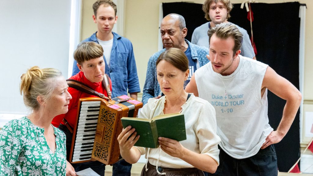 A group of actors rehearsing a play, one has an accordian, another is holding a book and all the others are looking over her shoulder. They look confused.
