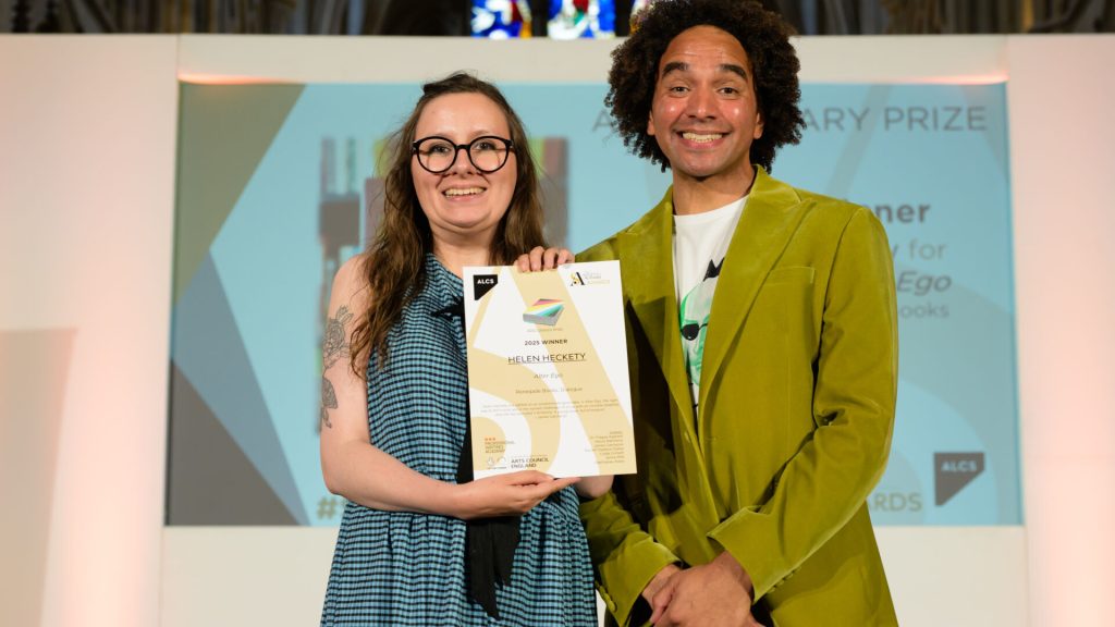 A smiling man and woman together on a stage. The woman is holding up a certificate.