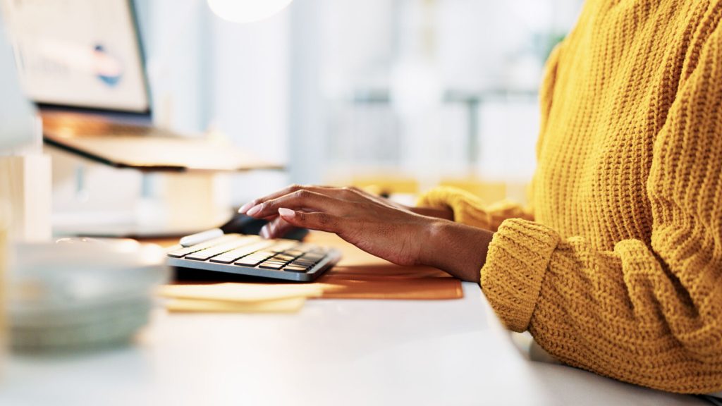 A person in a yellow jumper sitting at a laptop, you can just see their arms and hands.