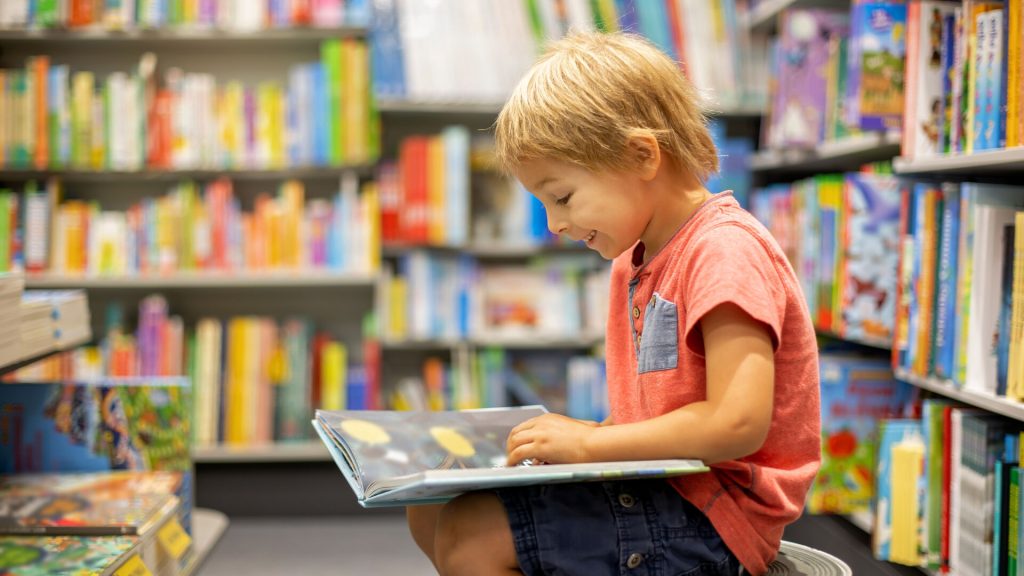 young boy happily reading, surrounded by books in a library or book shop.