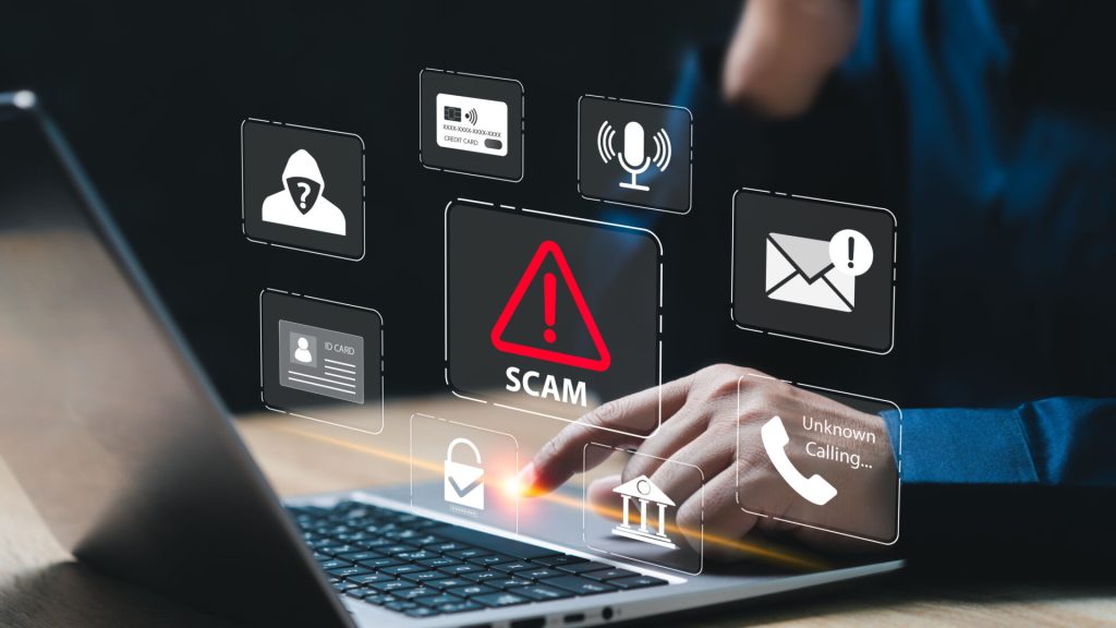 A person on a laptop with a number of icons floating in the air and red warning triangle with the word Scam in the middle