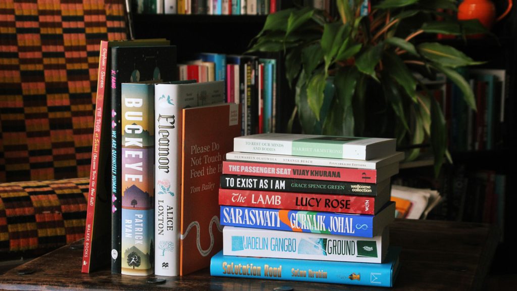 A stack of books set against a plant and a bookshelf
