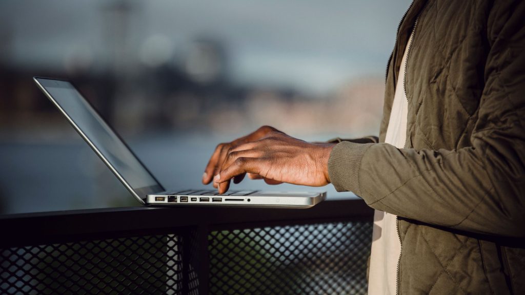 Side view of crop anonymous black male freelancer in casual wear using laptop to work on remote project while standing on open terrace